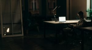 A man sits alone at his desk at night, looking at his phone